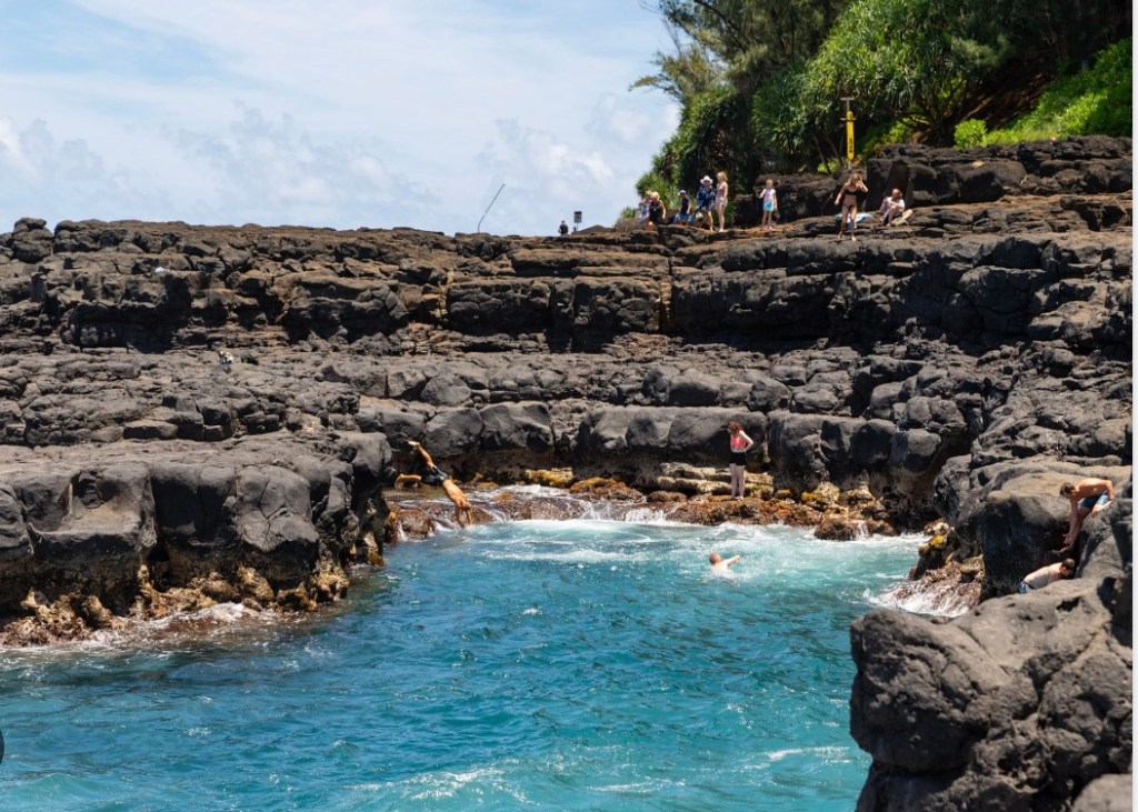 Swimmers and visitors on the black lava rock shelves surrounding Queen's Bath in Princeville on Kauai's North Shore, a natural tidal pool carved into volcanic rock with deep turquoise water on a sunny summer day.