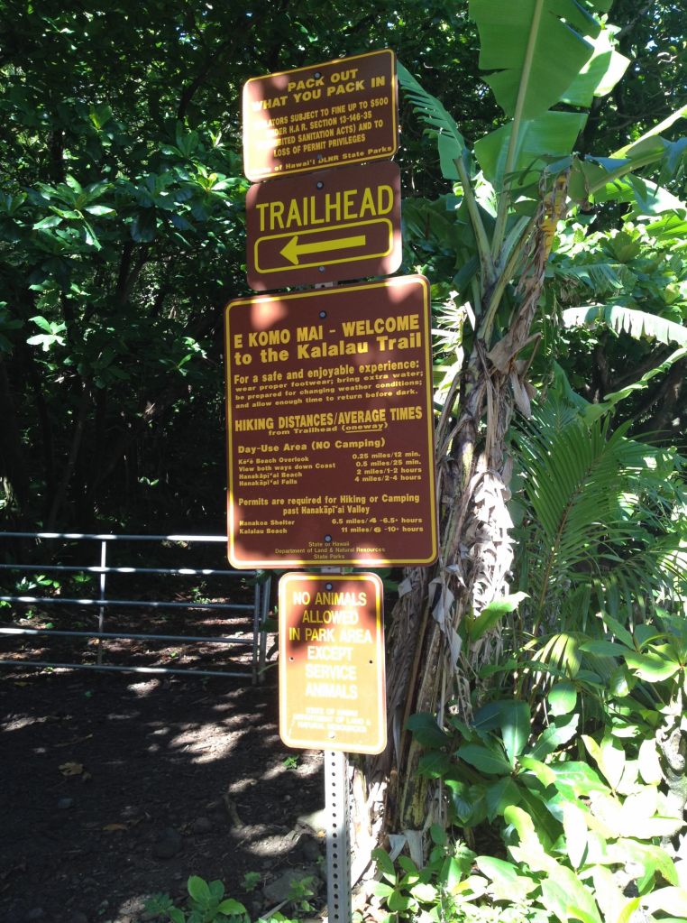 A brown metal sign marks the trailhead for the Kalalau trail with warnings to hikers.