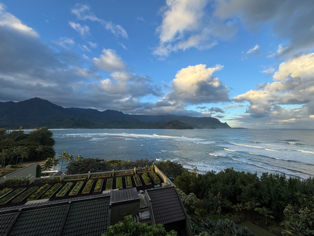 The view from the Walina Terrace at 1 Hanalei in Princeville, looking over the resort's rooftop garden toward Hanalei Bay, with surf lines rolling in and the green mountains and Makana peak rising along Kauai's North Shore under a dramatic morning sky.
