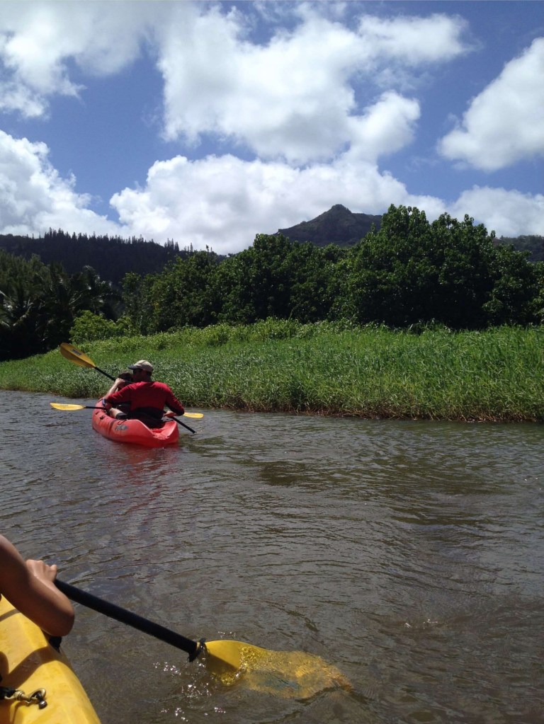 Kayakers paddling red and yellow kayaks on the calm Wailua River with Sleeping Giant mountain visible in distance