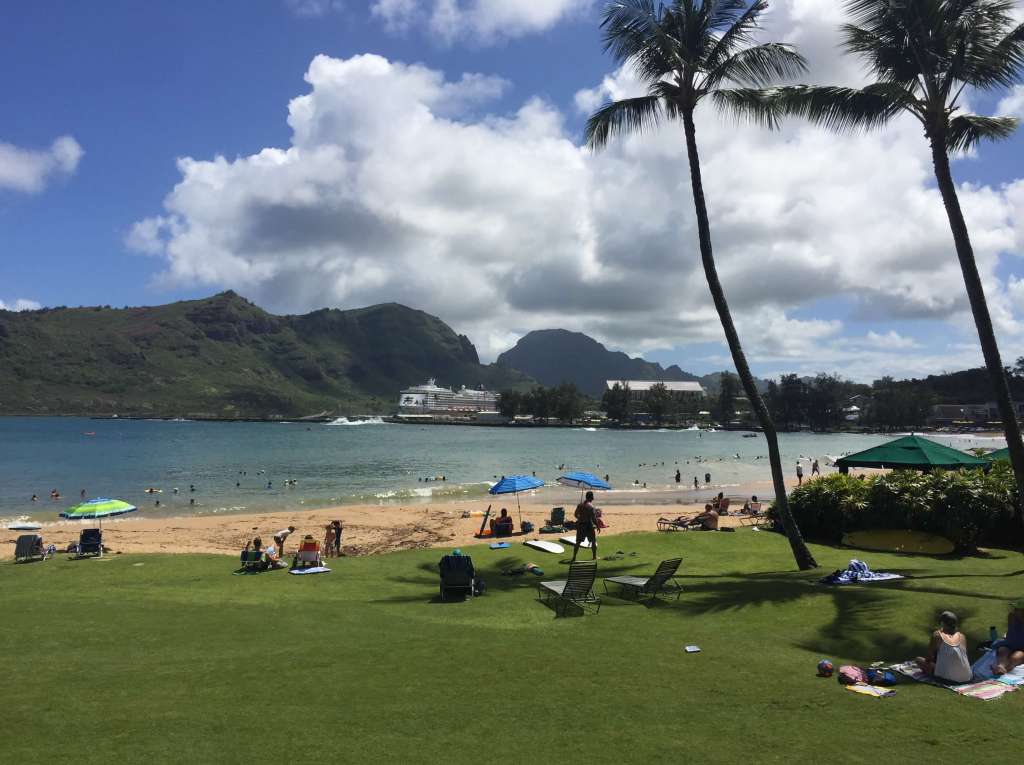 Green grasses and golden sands rest alongside the blue waters of Kalapaki Bay with green mountains and a cruise ship visible in the distance