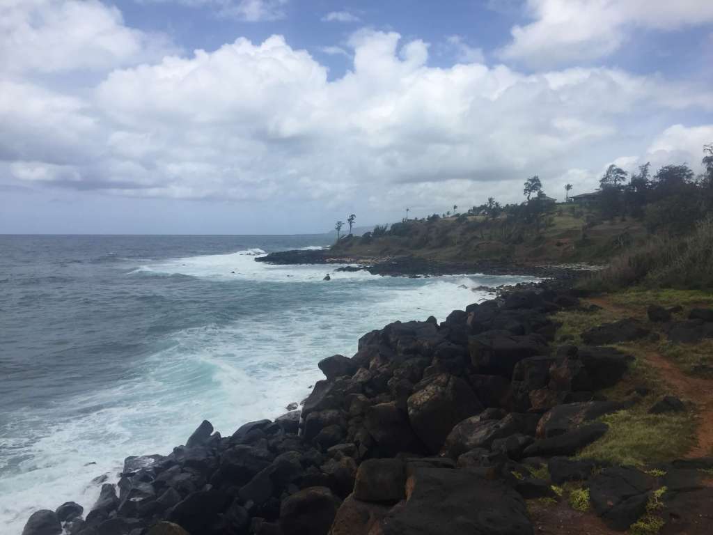 Rugged lava rock coastline on Kauai's East Shore, with turquoise waves crashing against black volcanic rocks and a green hillside with palm trees and a house visible in the distance under a partly cloudy sky.