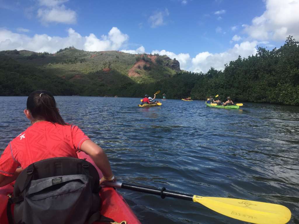Kayakers paddling red, green, and yellow kayaks on the calm Wailua River with jungle on both sides