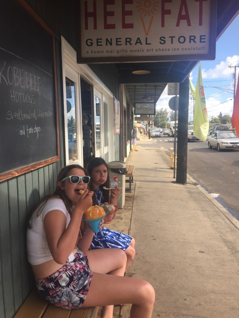 Shave ice from Hee Fat General Store in Kapa'a, Kauai's East Shore, enjoyed on the sidewalk bench outside the shop with the store's signature sign and Kapa'a's main street visible in the background.