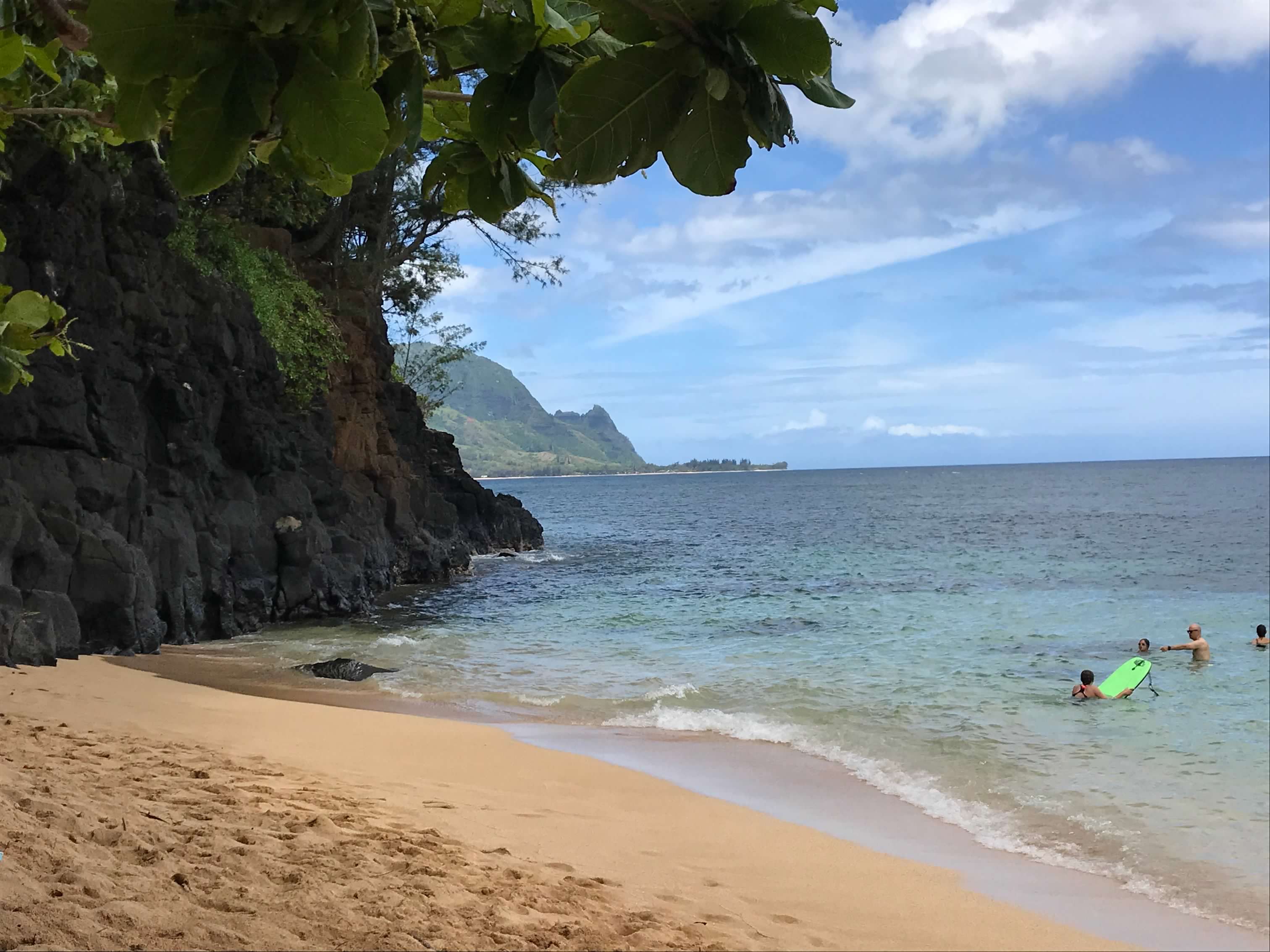 View from Hideaways Beach on Kauai's North Shore, showing a small secluded sandy cove with crystal-clear turquoise waters where snorkelers enjoy the calm conditions. The beach is framed by dramatic volcanic rock cliffs covered in moss and tropical vegetation, with large green leaves from coastal trees providing natural shade overhead. In the distance, the rugged Na Pali coastline extends into the Pacific Ocean under a partly cloudy sky. Several visitors can be seen swimming and snorkeling with flotation devices in the protected waters of this hidden gem beach.