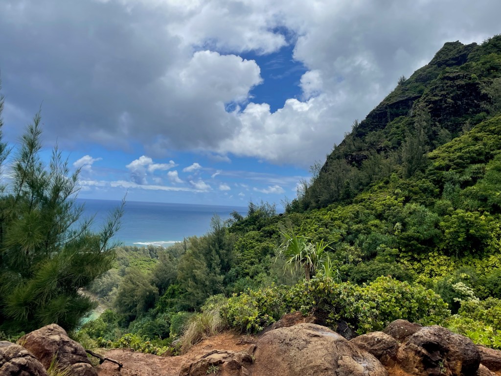 The lookout view over Ke'e Beach from the start of the Kalalau Trail on Kauai's North Shore — lush green cliffs, native vegetation, and turquoise ocean visible below under a partly cloudy summer sky.