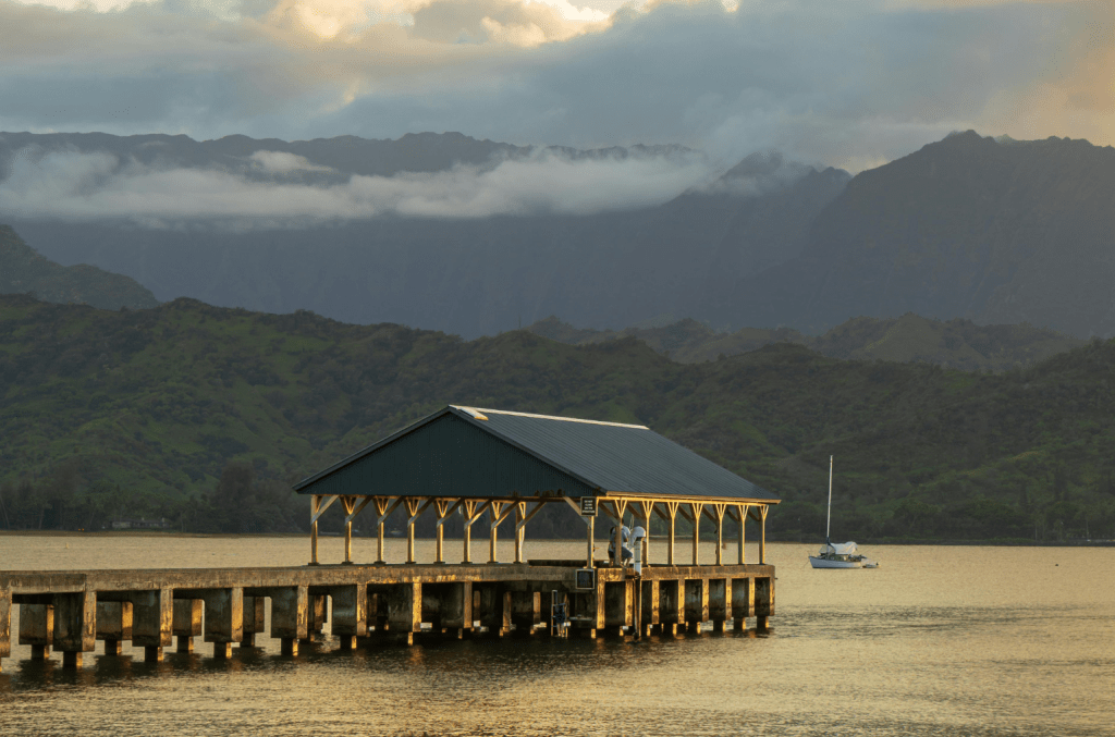 Hanalei Pier extending into the still waters of Hanalei Bay at dusk, with a sailboat anchored nearby and the green mountains of Kauai's North Shore disappearing into low clouds in the background.
