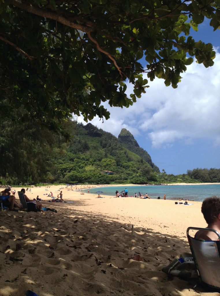 Beach goers enjoy the golden sands and clear, shallow waters at Tunnels Beach with the famed Bali Hai in the distance under lush tropical trees