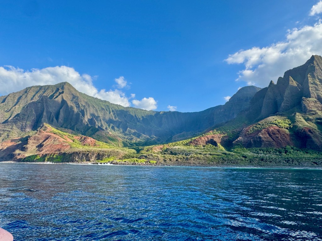 Dramatic view of Kalalau Valley from the ocean showing towering fluted cliffs, red volcanic rock formations, and lush green vegetation on Kauai's Na Pali Coast