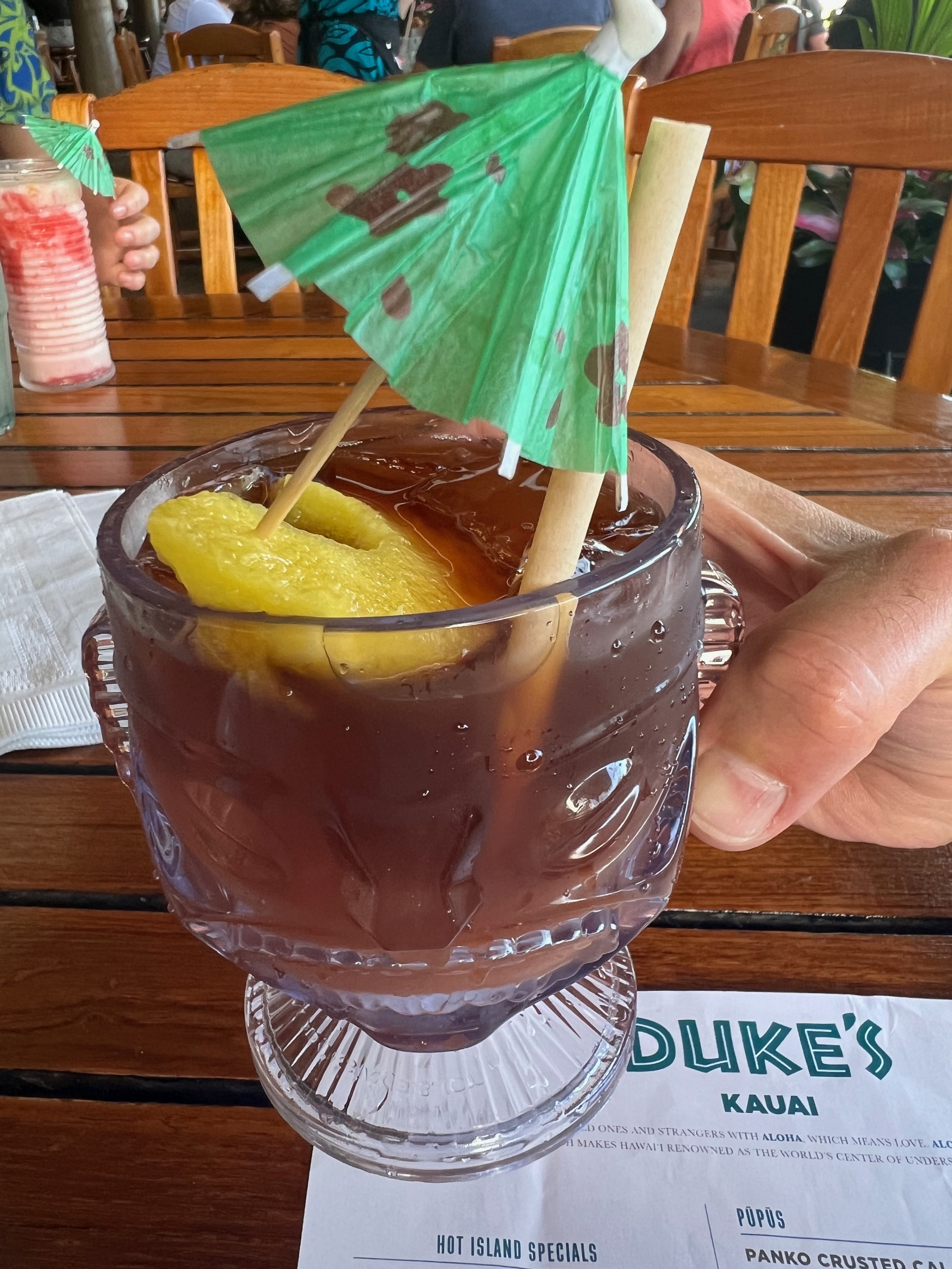 Classic mai tai cocktail in a clear tiki glass with a green umbrella sitting on a menu labeled Duke's in Lihue, Kauai
