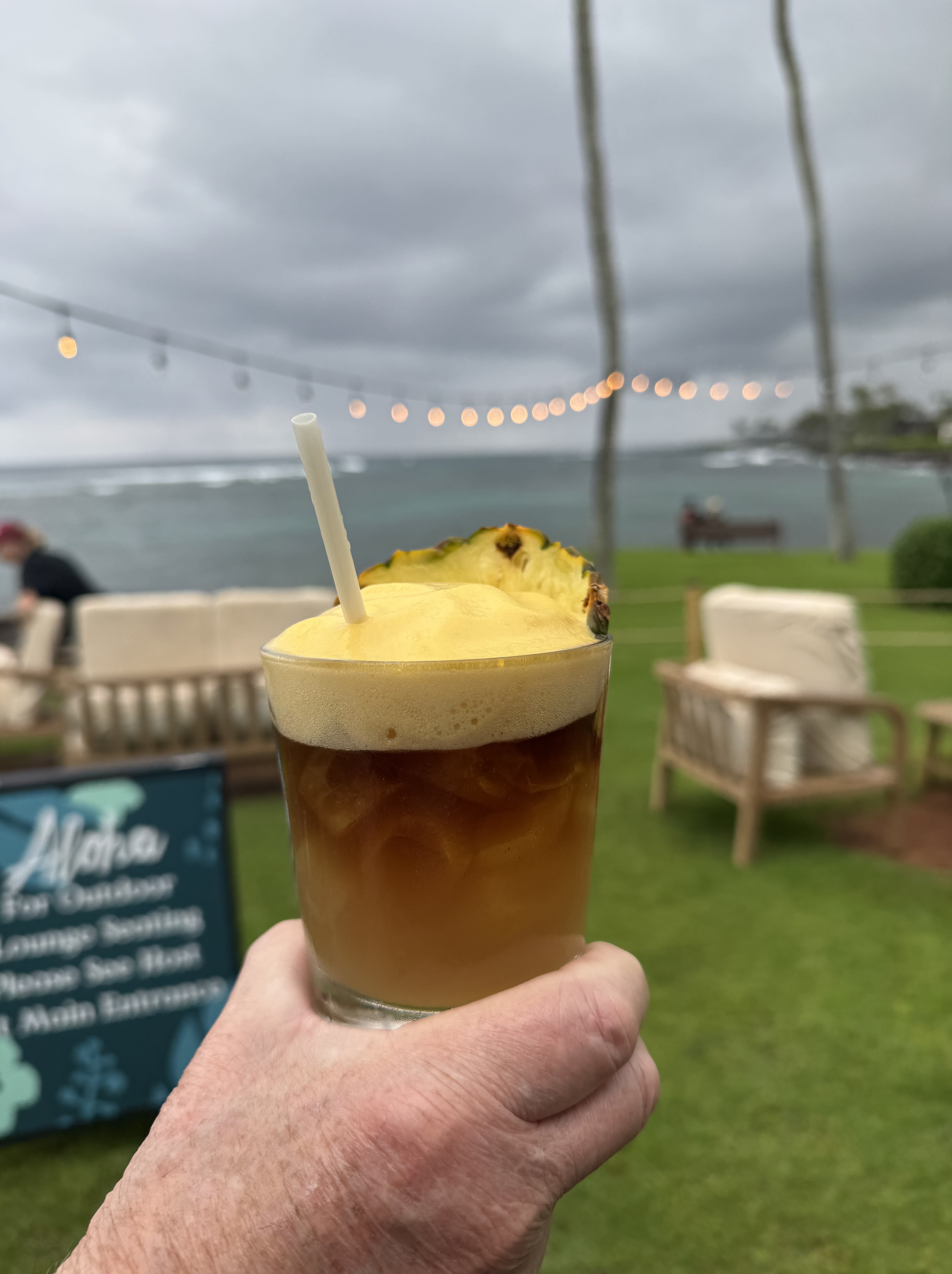 A hand holds a mai tai cocktail topped with passion fruit foam and garnished with a fresh pineapple wedge at The Beach House restaurant in Kauai. The drink is photographed against an overcast oceanfront view with string lights, green lawn, and wicker outdoor furniture visible in the background.