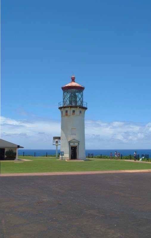 The historic Kilauea Lighthouse at Kilauea Point National Wildlife Refuge on Kauai's North Shore, built in 1913, with its distinctive red-capped tower rising against a bright blue sky and the Pacific Ocean stretching to the horizon behind it.
