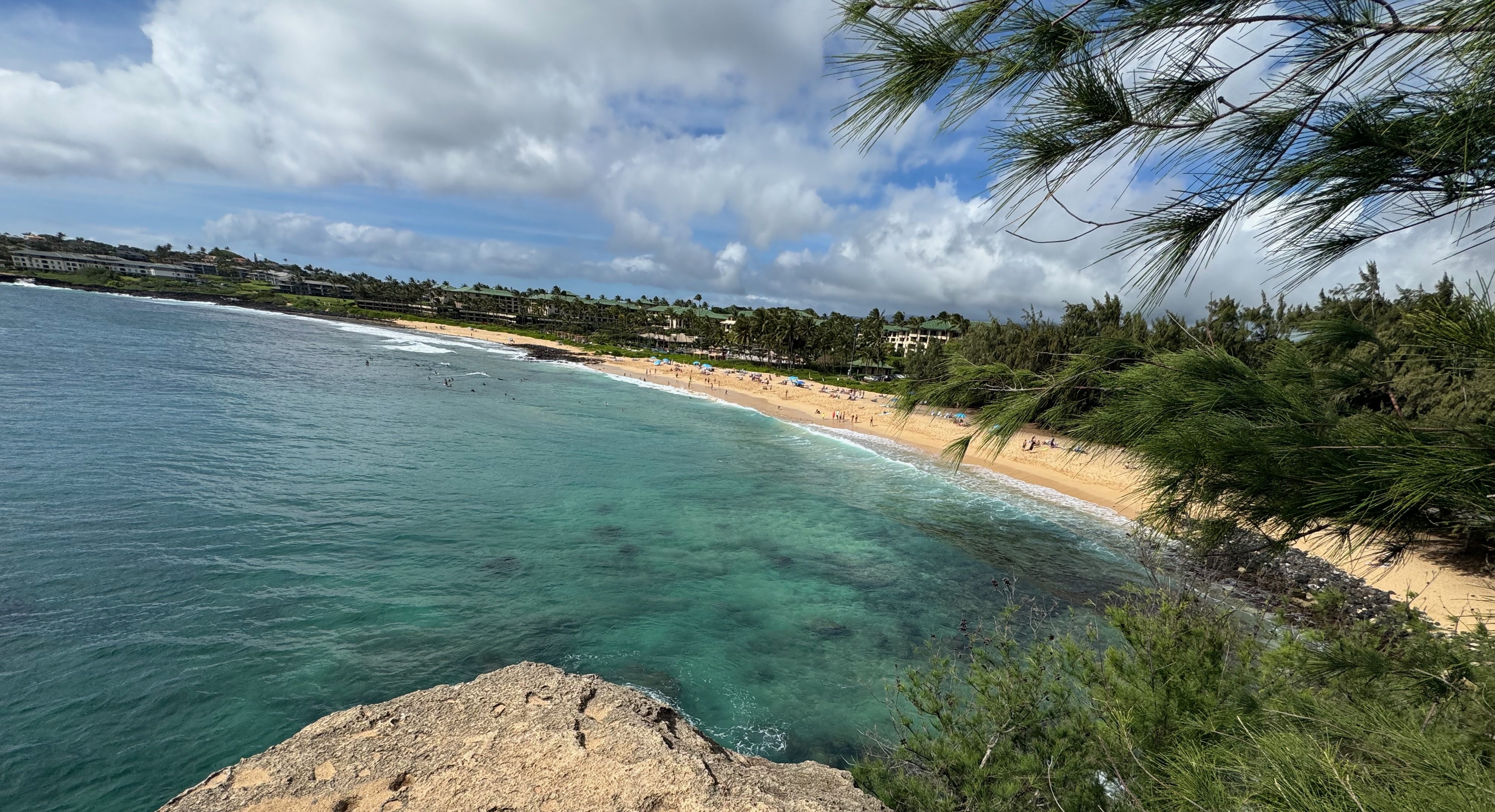 Shipwreck Beach on Kauai's South Shore viewed from the limestone cliffs above, showing the curved golden sand, turquoise water with visible reef, the Grand Hyatt resort in the background, and ironwood trees framing the foreground.