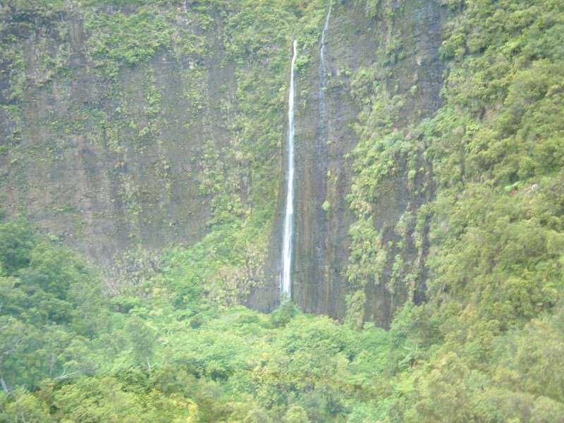 A tall waterfall cascading down the ancient basalt walls inside the crater of Mt. Wai'ale'ale on Kauai, photographed from a helicopter, showing the sheer moss-covered lava cliffs and dense jungle at the base of one of the wettest places on earth.